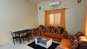 A living room with a sofa set, a dining table with chairs, a coffee table, and a ceiling fan. - Appartement meublé GOLF (Bamako)
