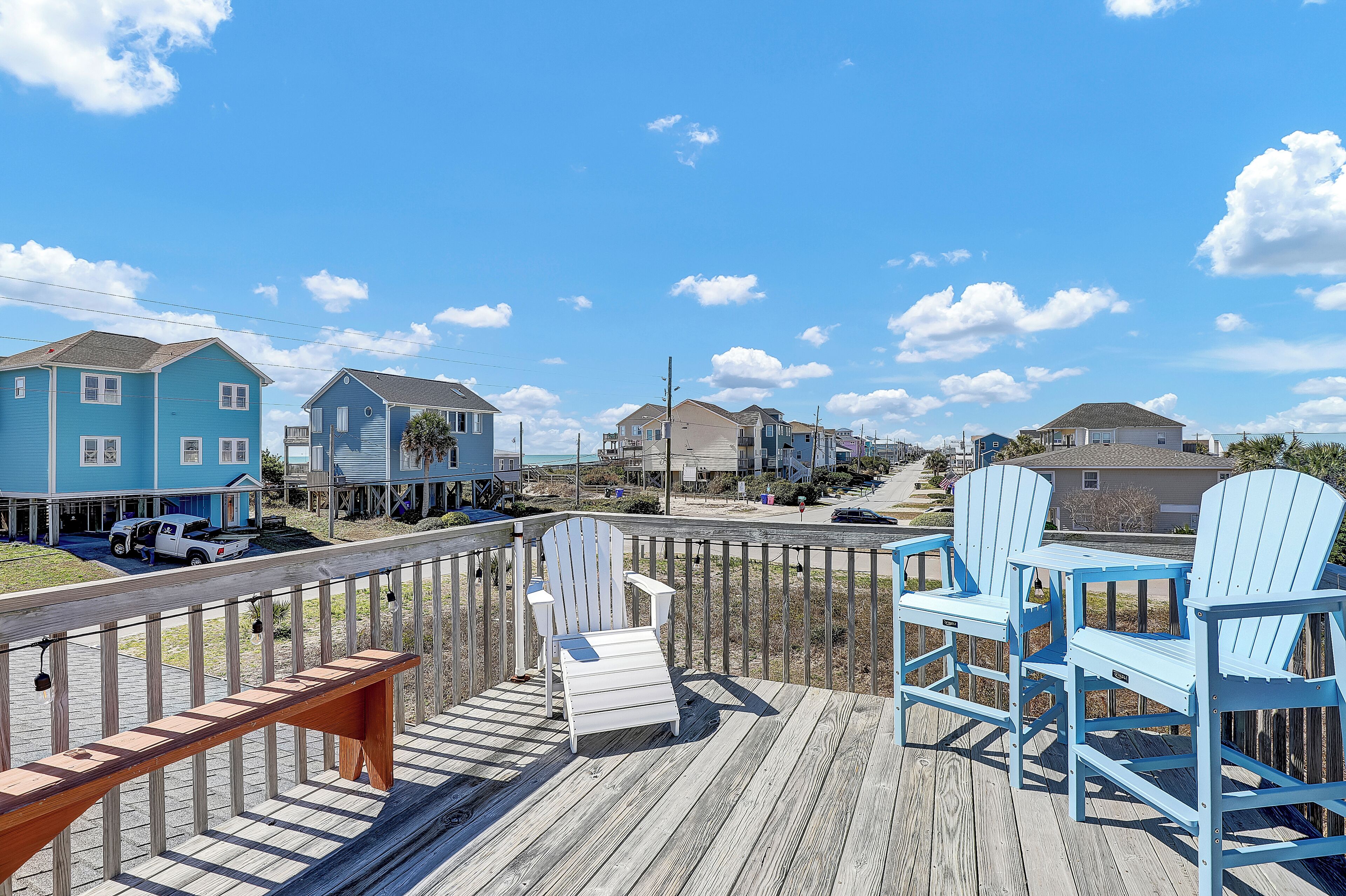 A wooden deck with blue Adirondack chairs and a bench, overlooking a residential area with houses and a road.