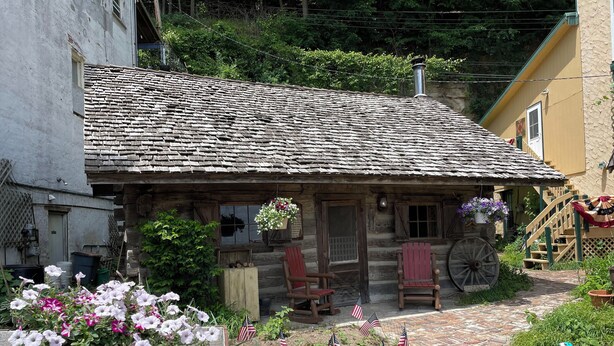 A rustic log cabin with a wooden porch, a wheelbarrow, and potted flowers. - Historic Sadie's Log Cabin (McGregor)