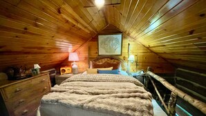 A cozy attic bedroom with a wooden sloped ceiling, a bed with a patterned bedspread, a dresser, a lamp, and a framed picture on the wall. - Historic Sadie's Log Cabin (McGregor)
