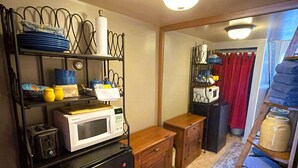 A compact kitchen with a microwave, toaster, and small refrigerator. There is a wooden cabinet and a metal shelf with dishes. A patterned rug is on the floor. - Cozy Alpine Retreat in Historic Downtown (McGregor)