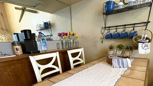 A kitchen with a long table, chairs, a coffee maker, a water dispenser, and a shelf with cups and a basket. - Cozy Alpine Retreat in Historic Downtown (McGregor)