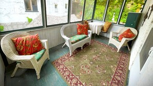 A sunroom with wicker chairs, a patterned rug, and large windows offering a view of a building and greenery outside.