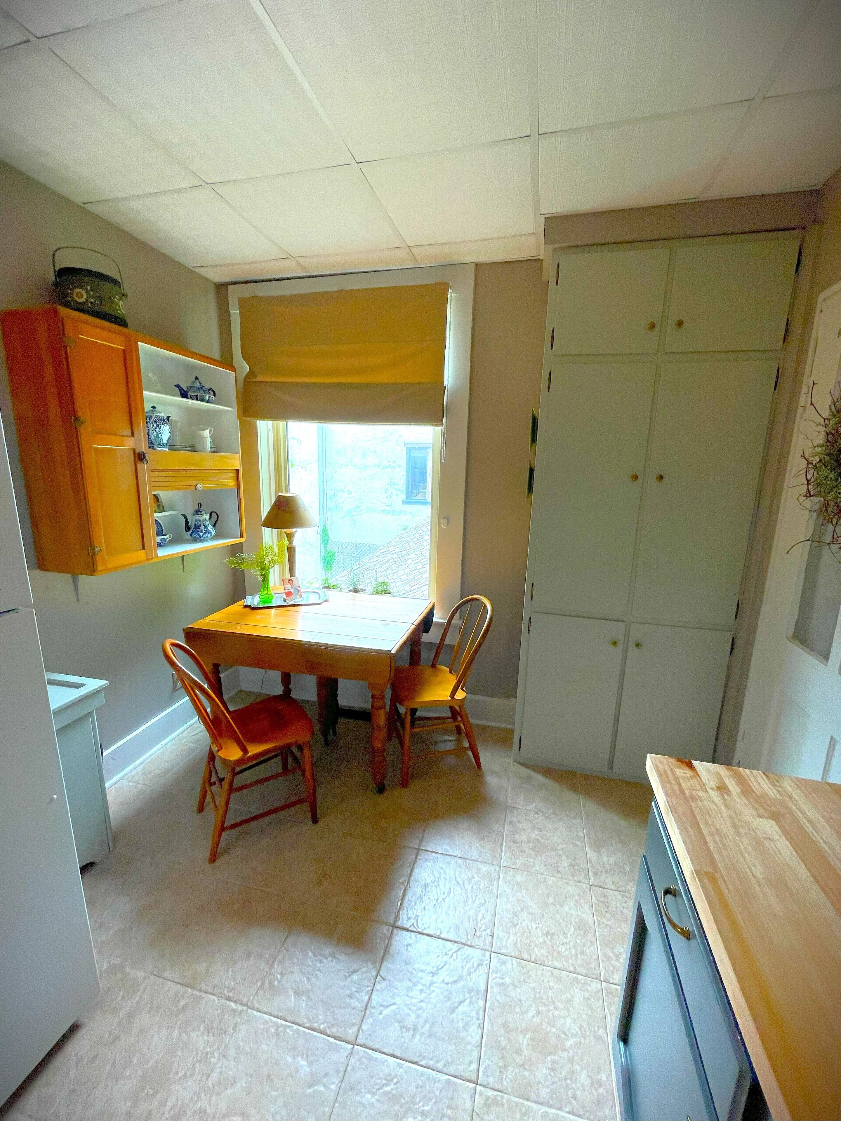A small kitchen with a dining table, two chairs, a cabinet with dishes, and a window with a yellow blind.