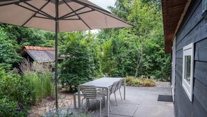 A patio with a table and chairs, surrounded by lush greenery and a house in the background. - Heivlinder Comfort 4p (Ermelo)