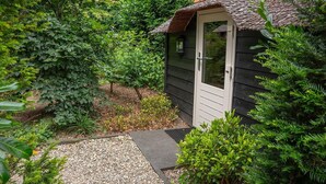 A small wooden shed with a door and a window, surrounded by dense greenery and a gravel path. - Heivlinder Comfort 4p (Ermelo)