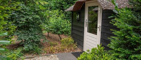 A small wooden shed with a door and a window, surrounded by dense greenery and a gravel path.