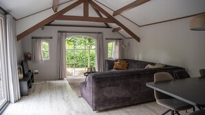A cozy room with a dark grey sofa, a dining table, and a TV on a stand. Large windows with curtains provide natural light and a view of greenery outside. - Heivlinder Comfort 4p (Ermelo)