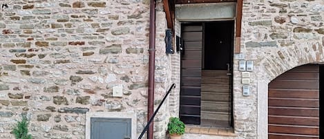 A stone building with a wooden door and steps leading to a dark interior. There are potted plants on the steps and a small window with shutters.