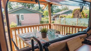A wooden deck with a table set for dining, a cushioned bench, and a view of a brick building and greenery. - Wander Lodge | 5 persons (Voorthuizen)