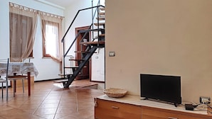 A living room with a tiled floor, a wooden TV stand, a flat-screen TV, a staircase with a wooden railing, and a dining area with a table and chairs.
