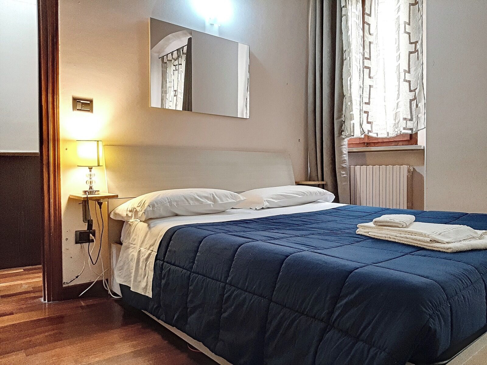 A hotel room with a bed, a nightstand with a lamp, a mirror, and a window with curtains.