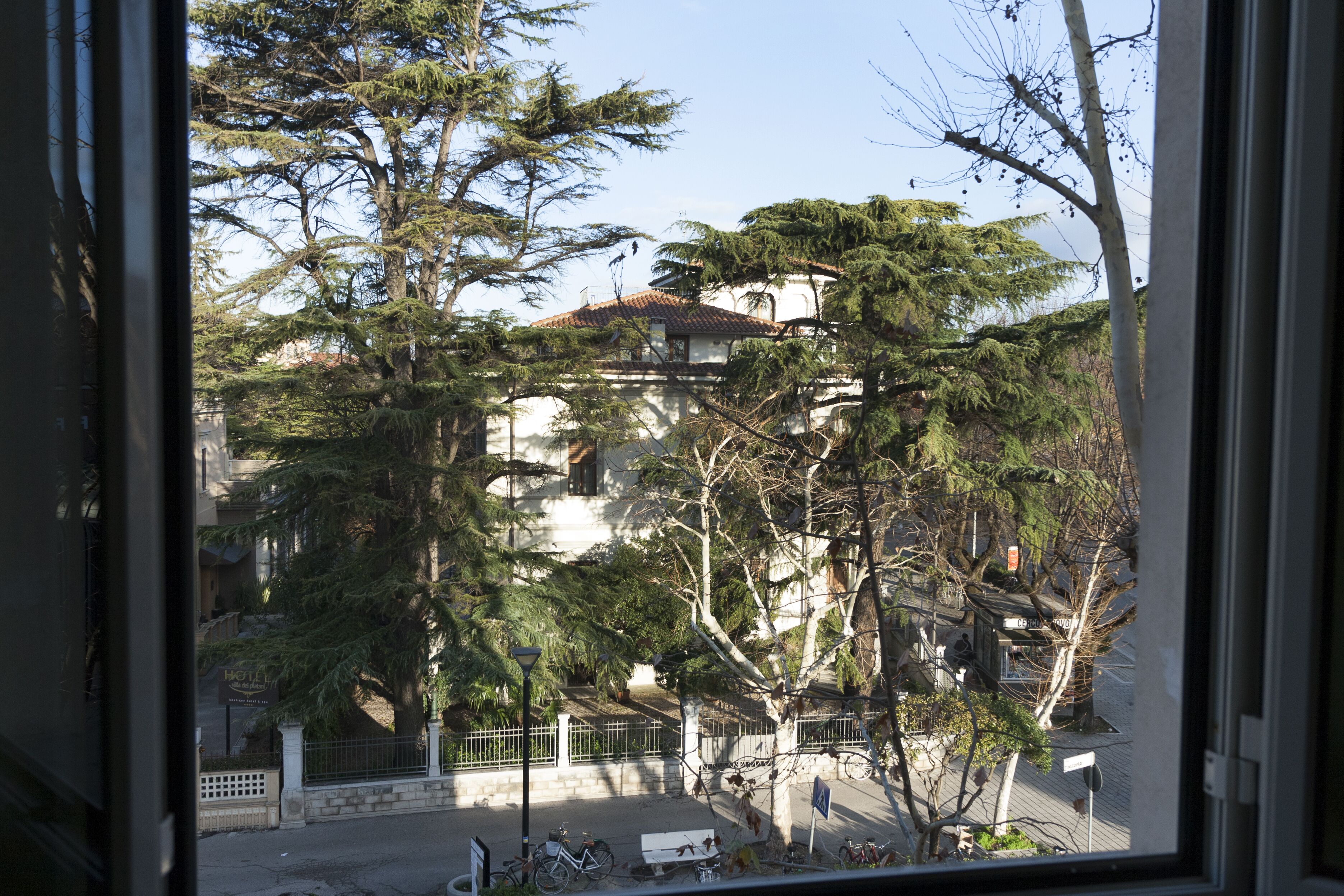 A view of a building with a tiled roof, surrounded by trees and a street with bicycles and a bench.