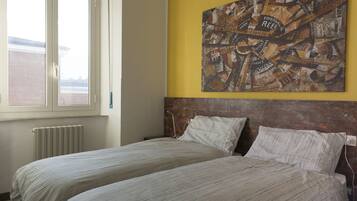 Une chambre avec deux lits, une fenêtre, un radiateur et un tableau représentant un paysage urbain accroché au mur.