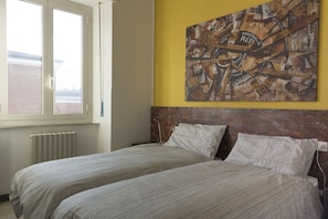 Une chambre avec deux lits, une fenêtre, un radiateur et un tableau représentant un paysage urbain accroché au mur.