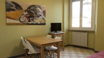 A room with a wooden table and white chairs, a television on a stand, a radiator, and a window with blinds.