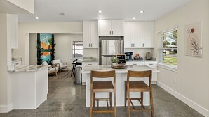 A modern kitchen with white cabinets, a stainless steel refrigerator, and a coffee bar. There are two wooden bar stools and a dining table. - Family-Friendly 3BR | Downtown & Lake Hollingsworth (Lakeland)