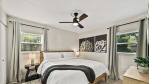 A bedroom with a bed, a ceiling fan, a nightstand, a lamp, a window with curtains, and a wall with three flower paintings. - Family-Friendly 3BR | Downtown & Lake Hollingsworth (Lakeland)