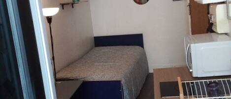A small, compact room with a bed, a desk, and a microwave. There are shelves with decorative items and a wall-mounted mirror.
