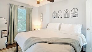 Een slaapkamer met een bed, een plafondventilator, een raam met gordijnen en wanddecoratie.