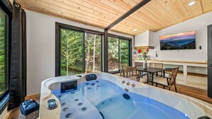 Un spa moderno con un amplio jacuzzi, techo de madera, ventanas grandes y una zona de comedor con mesa y sillas.