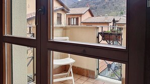 Un balcó amb una cadira blanca, una taula de metall i vista cap a cases properes i una muntanya.