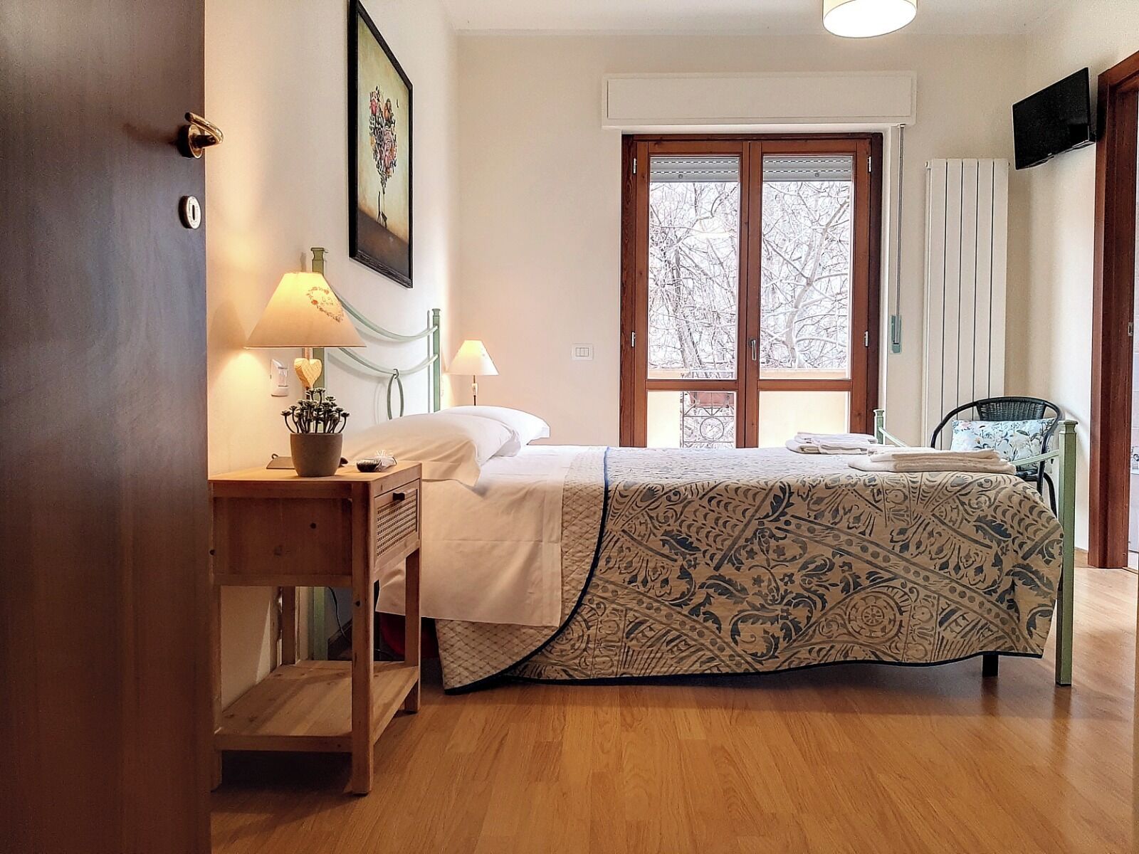 A bedroom with a bed, bedside lamps, a wooden nightstand, a chair, and a window with a view of trees.
