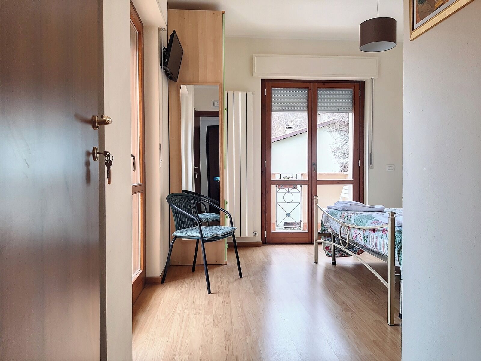 Ein schmales Entree mit Holzboden, einem Sessel, einem Bett mit Steppdecke und einer Tür mit Fenster.