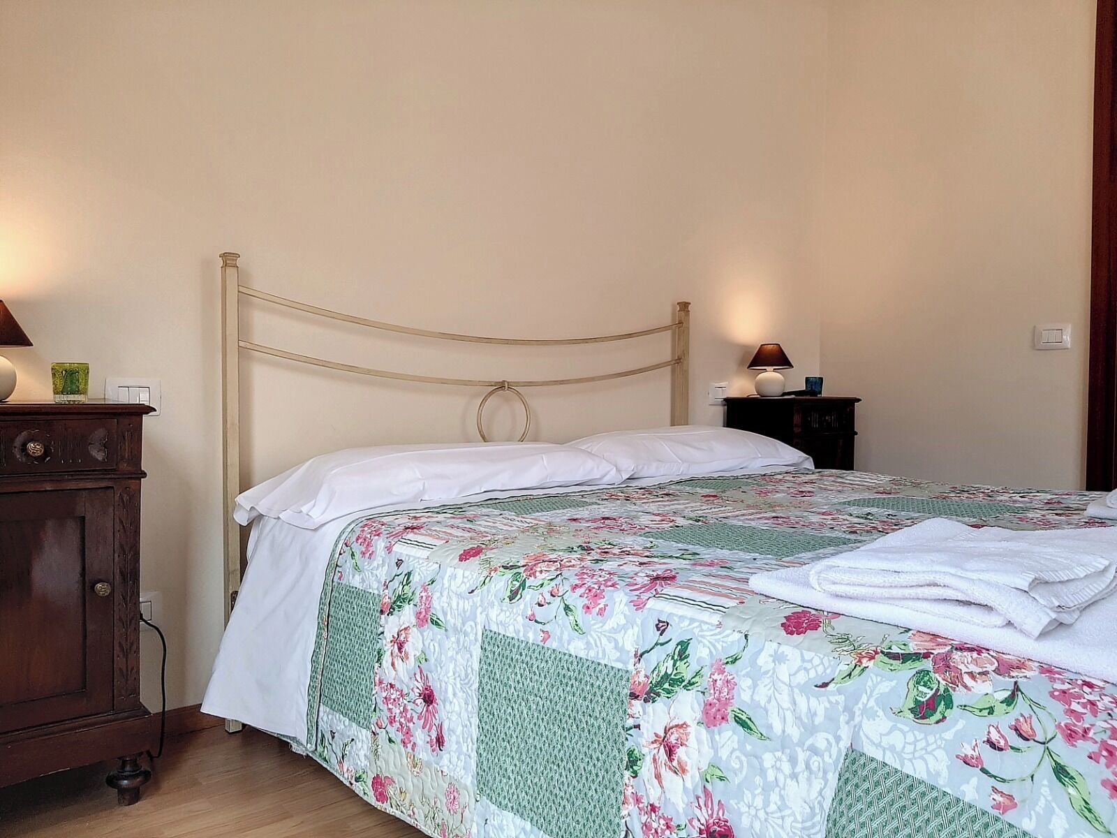 Un lit bien fait, recouvert d’une courtepointe à motifs floraux, une table de chevet en bois avec une lampe, et une commode en bois avec un miroir.
