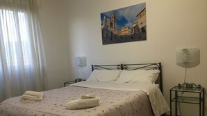 A neatly made bed with a floral bedspread and two folded towels, a painting of a town square on the wall, a bedside lamp, and a small table with a lamp. - Via Oberdan 113 by Ospitaci (Foligno)