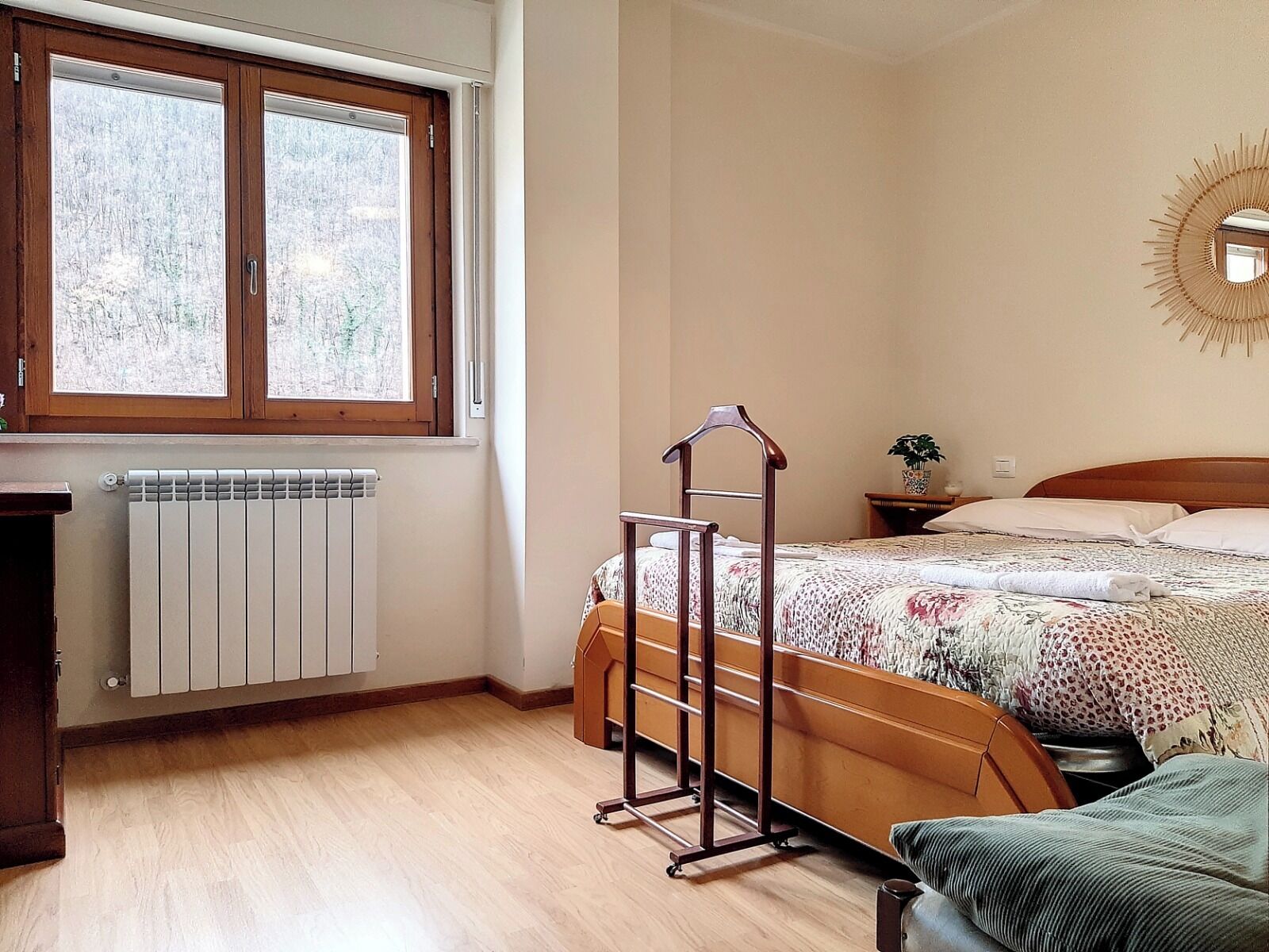 Une chambre avec un lit en bois, un radiateur, une fenêtre donnant sur des arbres et une table de chevet avec une plante.