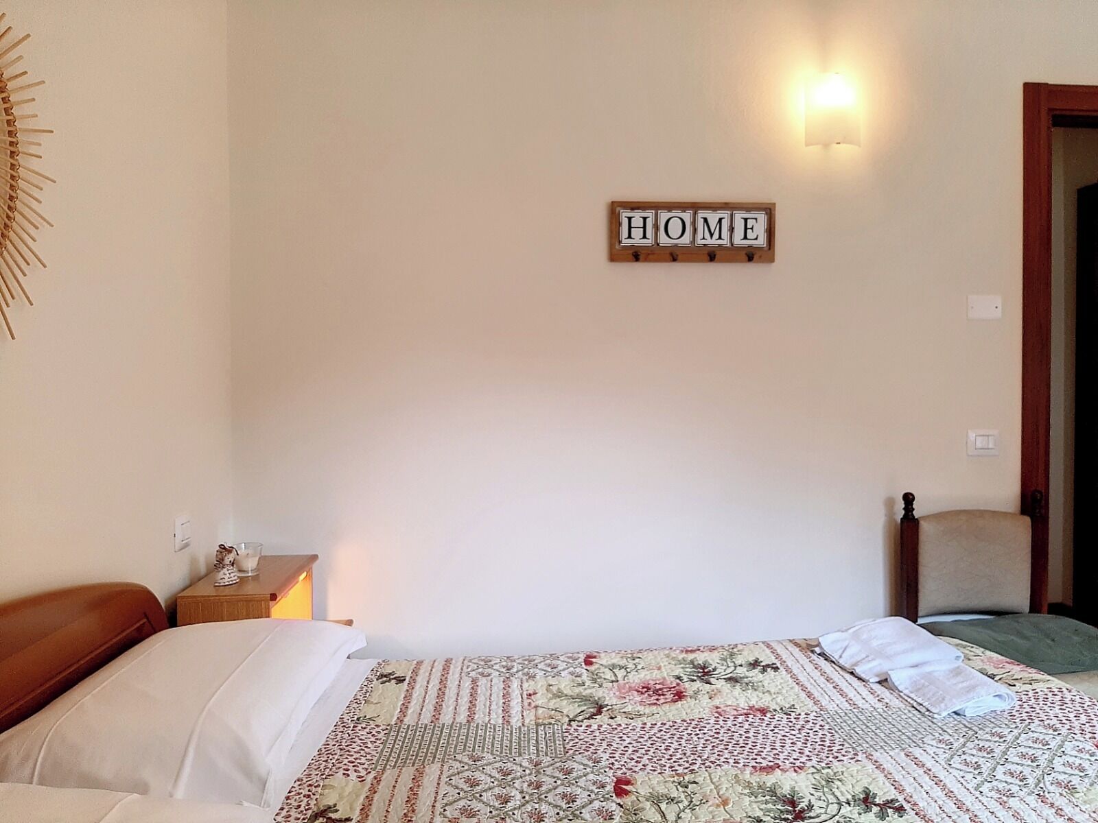 A bedroom with a bed, a bedside table with a lamp, a wall-mounted sign reading "HOME", and a chair in the corner.