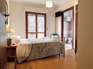 A bedroom with a bed, bedside table, lamp, and a view of the outside through a window.