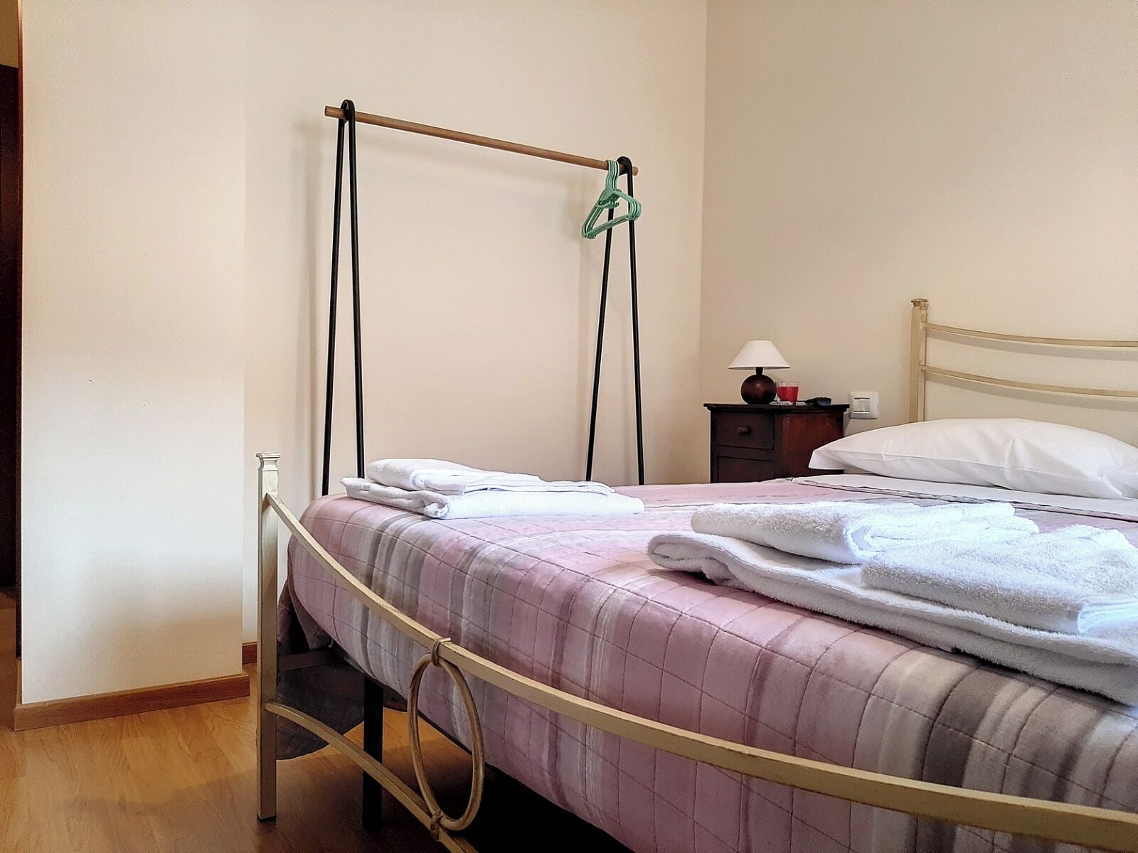 A neatly made bed with a pink quilt, a bedside table with a lamp, and a hanging towel rack.