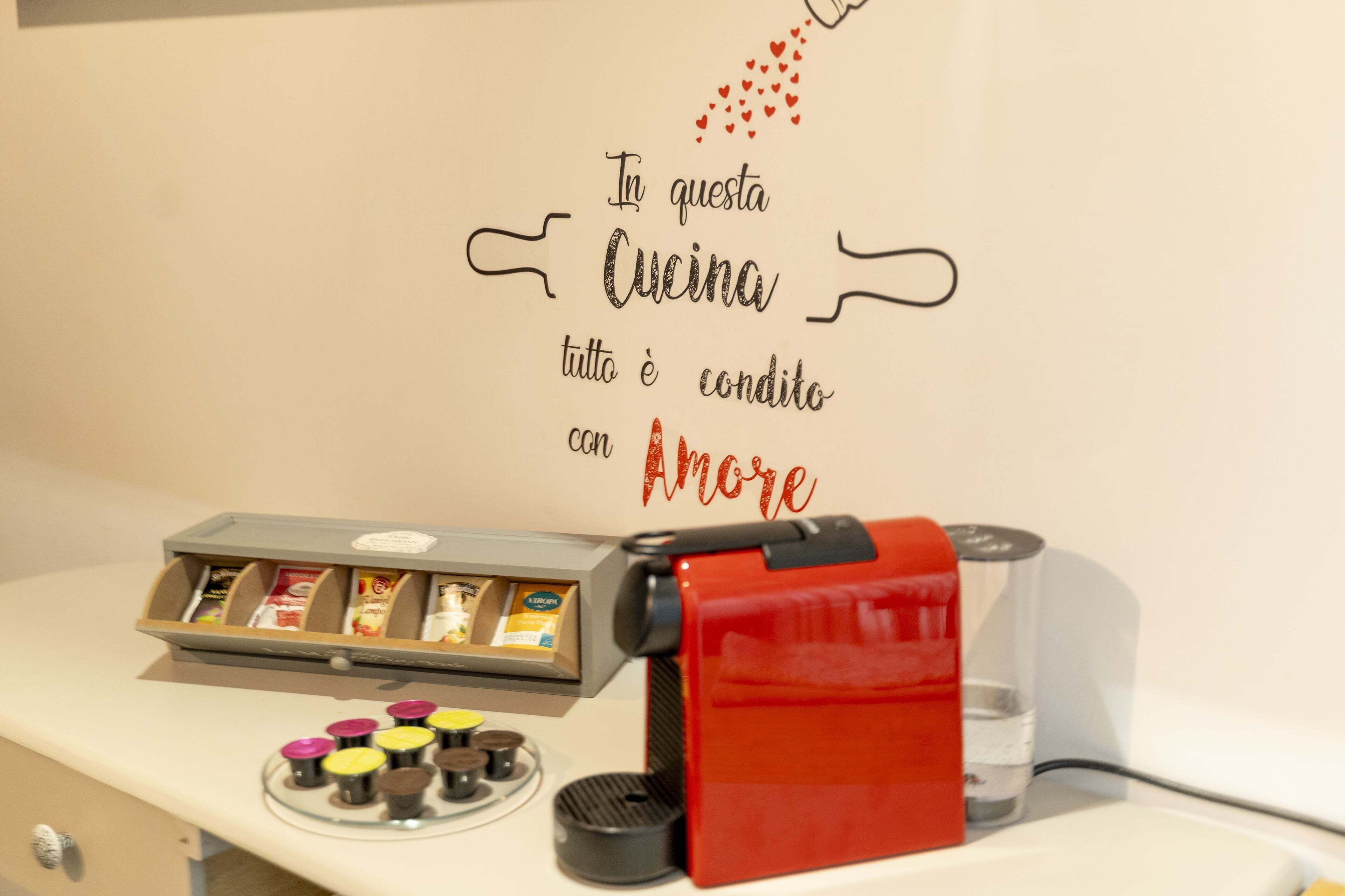 Una macchina da caffè e un vassoio di cialde per caffè su un bancone, con un cartello appeso al muro che recita: "In questa cucina tutto è condotto con Amore".