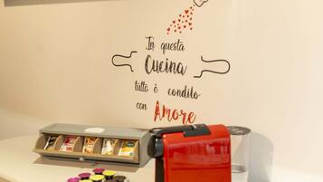Una macchina da caffè e un vassoio di cialde per caffè su un bancone, con un cartello appeso al muro che recita: "In questa cucina tutto è condotto con Amore".