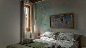 Une chambre à coucher avec un lit, des oreillers, un tableau, une fenêtre avec des rideaux et une lampe.
