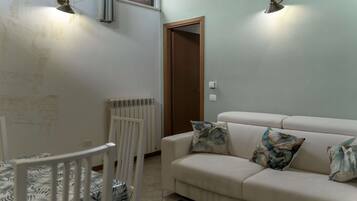 Un salon avec un canapé beige, deux coussins à motifs, un radiateur blanc, une chaise blanche et une lampe fixée au mur.