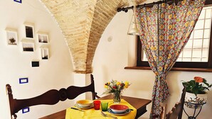 A dining area with a wooden table, chairs, and a yellow tablecloth. There is a window with a curtain, a potted plant, and a wall with framed pictures.