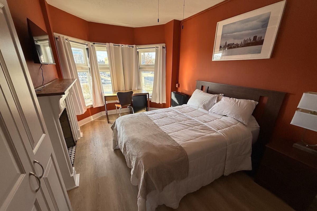 A hotel room with a large bed, a desk with a chair, a radiator, and a cityscape picture on the wall.