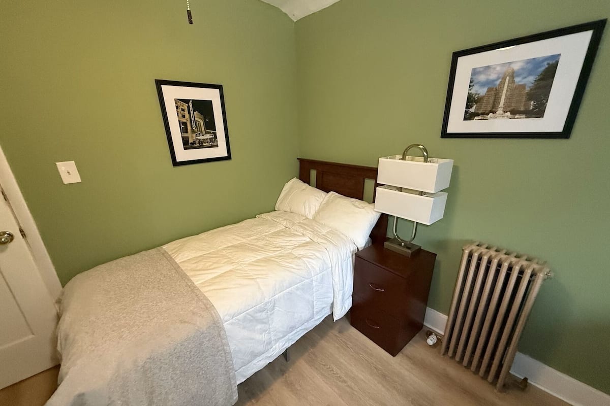 A neatly made bed with a white comforter and pillows, a bedside table with a lamp, a framed picture on the wall, and a radiator.
