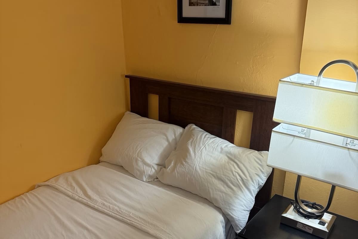 A single bed with white bedding, a wooden headboard, a framed picture on the wall, and a bedside table with a lamp.