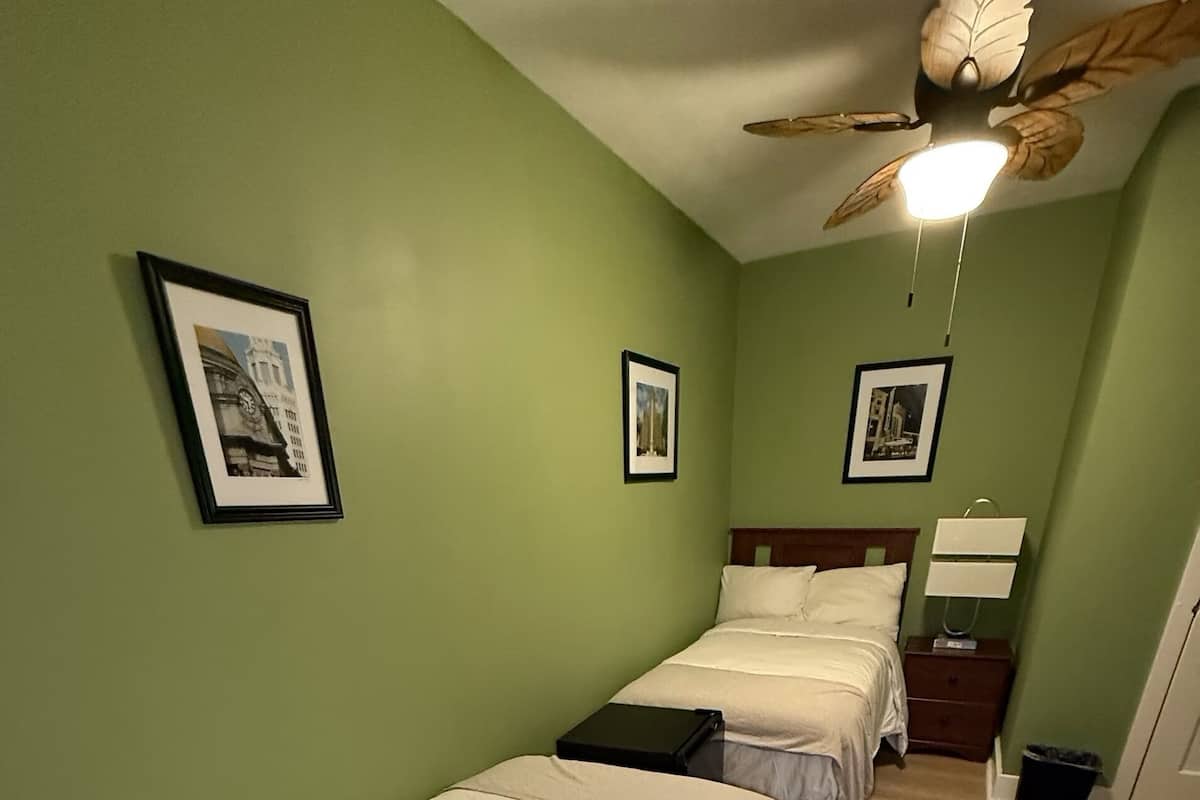 A room with two single beds, a ceiling fan, and framed pictures on the walls.