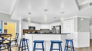 A modern kitchen with white cabinetry, a central island with stools, and a dining area with a table and chairs. - ☀️Panoramic Gulf and Pool Views-Steps to Beach-Calypso 1-1709☀️ (Panama City Beach)