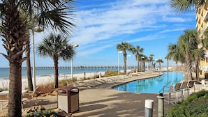 A pool area with lounge chairs, palm trees, and a view of the beach and pier. - ☀️Panoramic Gulf and Pool Views-Steps to Beach-Calypso 1-1709☀️ (Panama City Beach)