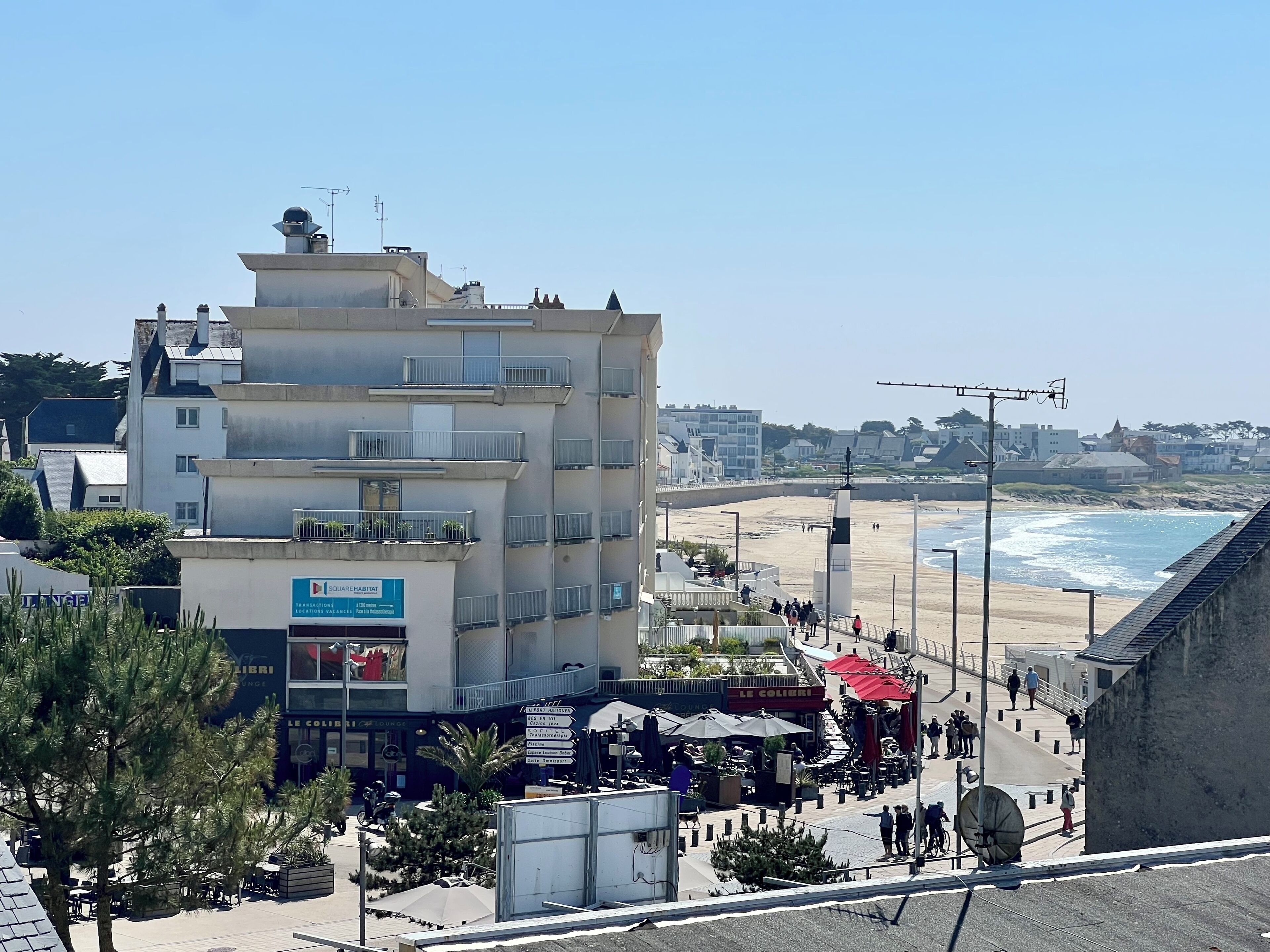 A multi-story building with balconies, a beach in the background, and a clear sky.