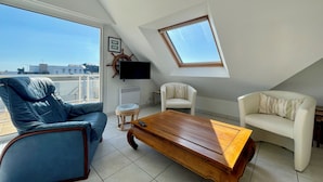 A modern living room with a blue leather sofa, a wooden coffee table, and a white armchair. A TV is mounted on the wall, and a ship's wheel decoration is visible.