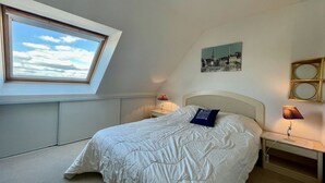 A bedroom with a skylight, a bed with a blue pillow, a nightstand with a lamp, and a wall-mounted shelf.