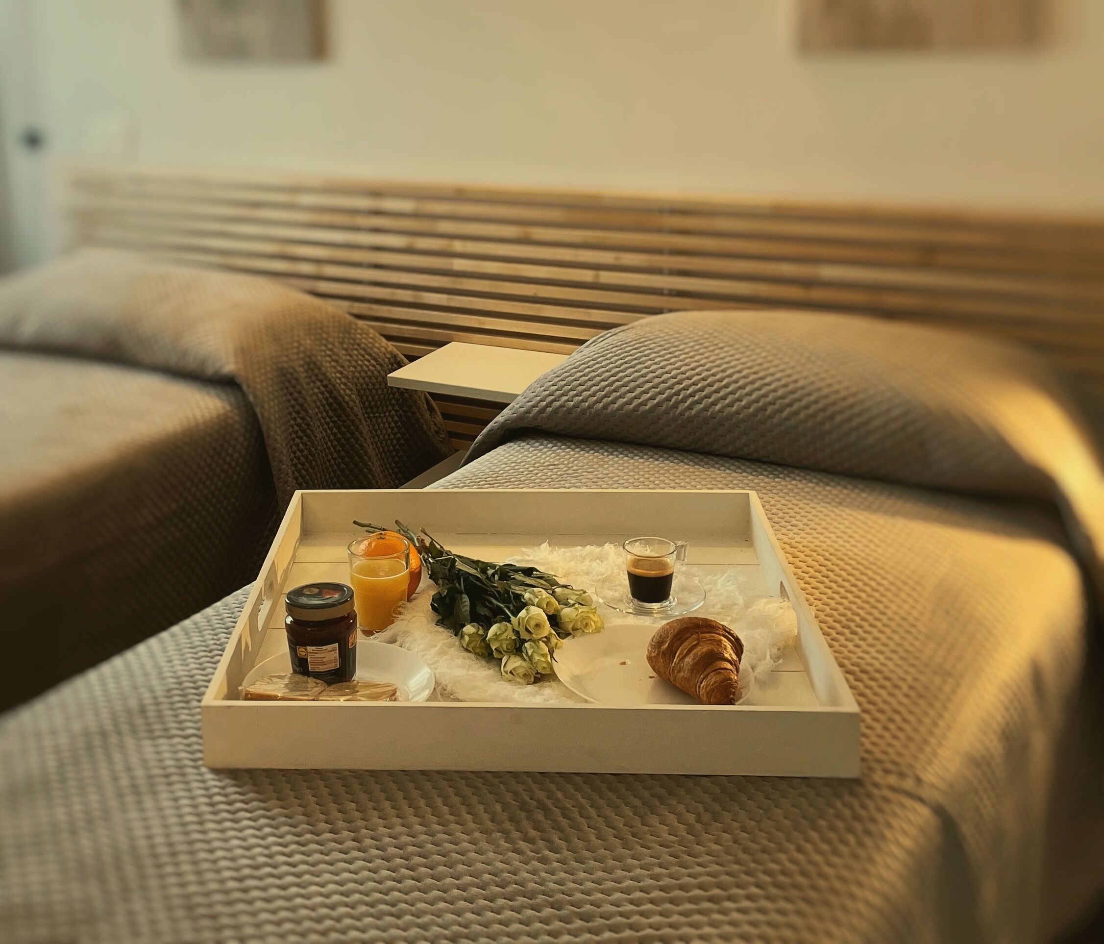 Un plateau de petit-déjeuner sur un lit, avec une bouteille de jus, un verre de jus d’orange, un croissant et un petit bouquet de fleurs.