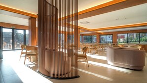 A spacious lobby with a modern design, featuring a large seating area with sofas and chairs, a wooden decorative column, and large windows offering views of the outdoors. - Golden Tulip Canela OFICIAL (Canela)